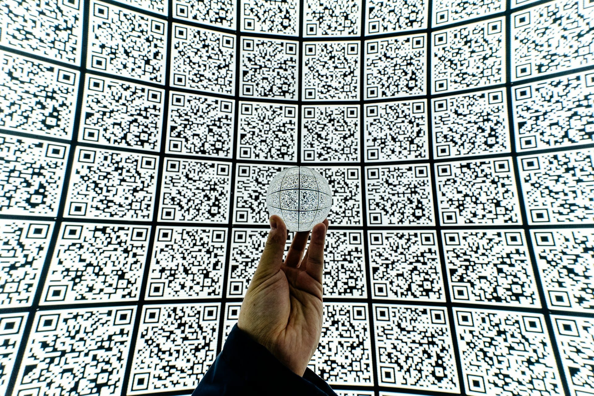 A large wall of QR codes. In front, a hand holds a clear sphere, reflecting the QR codes