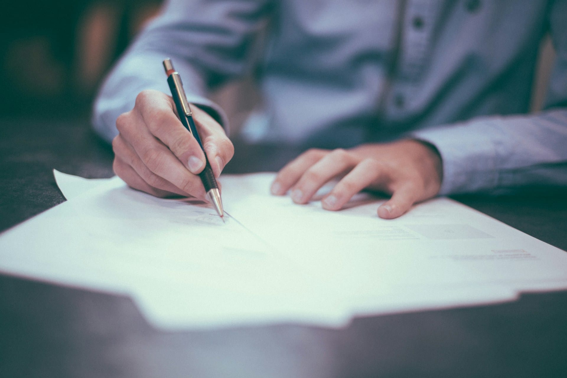 A close-up of a white paper spread on a table, as a person writes on a page with a black pen