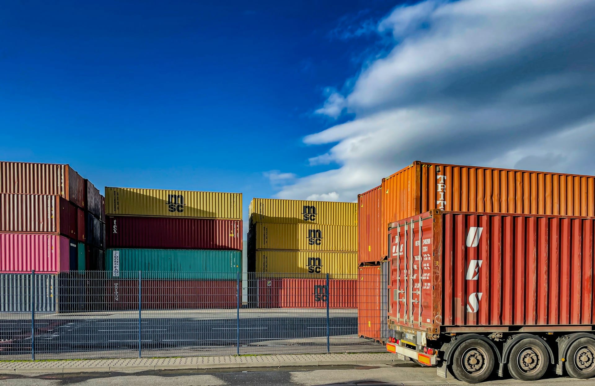 An image of large, stacked shipping containers in a shipping yard. An orange truck is parked to the right.